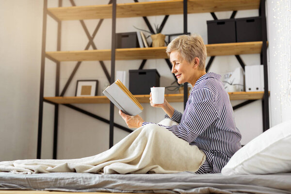  beautiful elderly woman in pajamas sitting in bed with a book and a mug