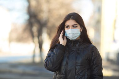 Sokakta elinde telefonuyla tıbbi maske takan genç ve güzel bir kadın.