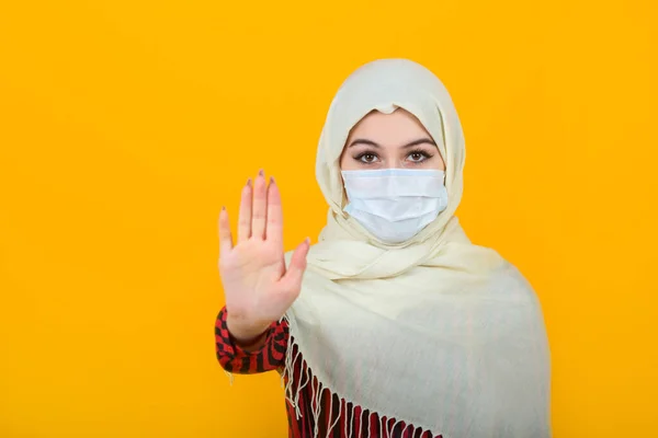 手のジェスチャーでイスラム教徒のショールの黄色の背景に医療用マスクの美しい若い女性 — ストック写真