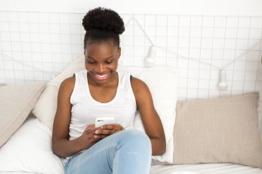 Güzel, genç, Afrikalı bir kadın telefonuyla yatakta oturuyor.