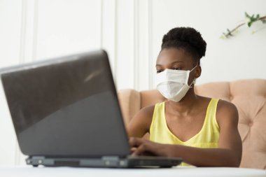 Güzel, genç, Afrikalı bir kadın bir sandalyede oturuyor. Elinde laptopuyla tıbbi bir maske var.