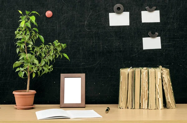 Teacher or student desk table. Education background. Education concept. Stacked books, copybook, book, green plant tree, photo frame and note paper on blackboard background.