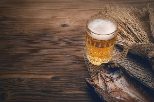 Foam beer in a mug, dried fish stockfish and rye ears wrapped in burlap cloth on a burnt wooden table background with copy space.