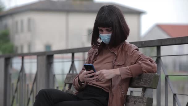 Tıbbi maskeli genç bir kadın parkta akıllı telefonuna bakıyor. — Stok video