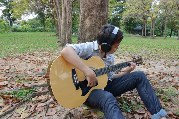 Retrato de jovem feliz tocando guitarra acústica e inclinando uma árvore no parque ao ar livre . — Fotografia de Stock