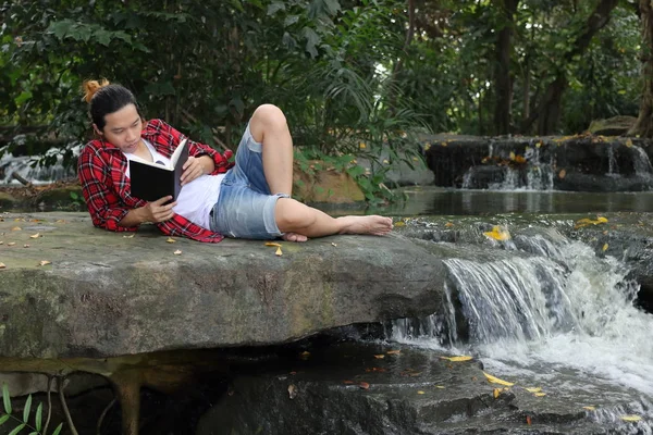 Retrato de un joven hombre relajado en camisa roja tirado en el suelo y leyendo un libro en hermoso fondo de la naturaleza . — Foto de Stock