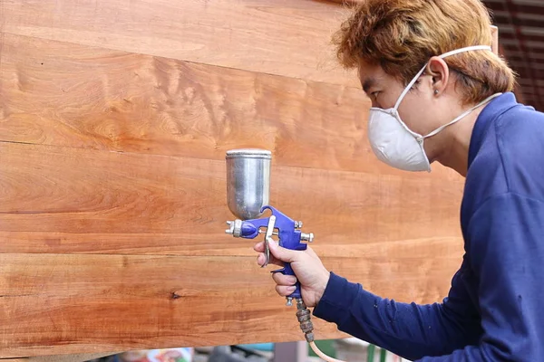 Selektiver Fokus auf die Hände eines jungen asiatischen Arbeiters mit Schutzmaske, der in der heimischen Werkstatt ein Stück Holz mit einer Spritzpistole bemalt. geringe Schärfentiefe. — Stockfoto