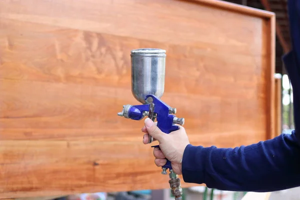 Manos de trabajador industrial aplicando pistola de pintura en aerosol con un mobiliario de madera el fondo del taller . —  Fotos de Stock