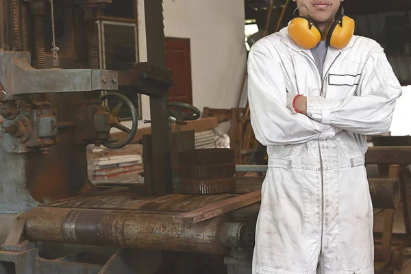 Porträt eines selbstbewussten jungen Arbeiters in weißer Uniform, der mit Bandsägen-Hintergrund die Arme über der Brust verschränkt. — Stockfoto