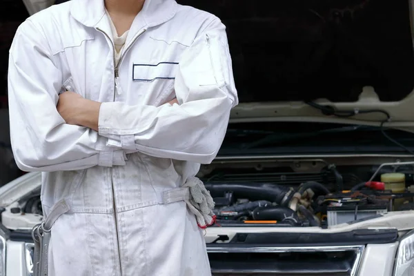 Ritratto di giovane meccanico fiducioso in uniforme con braccia incrociate e in piedi contro l'auto in cappa aperta al garage di riparazione . — Foto Stock