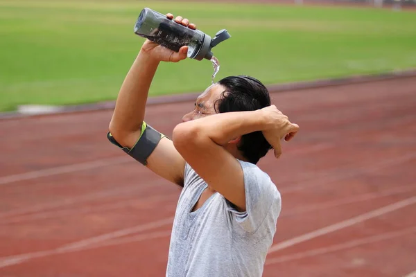 Young Asian fitness man splashing water on his face after run in track of stadium.