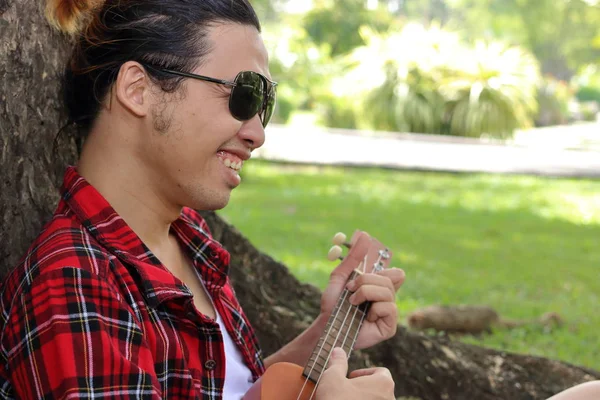 Jovem tocando ukulele no parque ao ar livre. Ele está sorrindo. — Fotografia de Stock