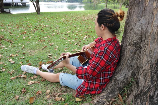 Zadní pohled na uvolněné mladíka v červené košili, opřený o strom a hrál na akustickou kytaru v krásném venkovním parku. — Stock fotografie