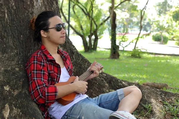 Portrét mladého muže, uvolněně leží a hrát na ukulele na veřejné venkovní park. — Stock fotografie