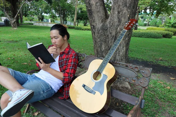 Porträtt av ung asiatisk man i röd skjorta en bok på bänken mot i vackra Utomhus park. — Stockfoto