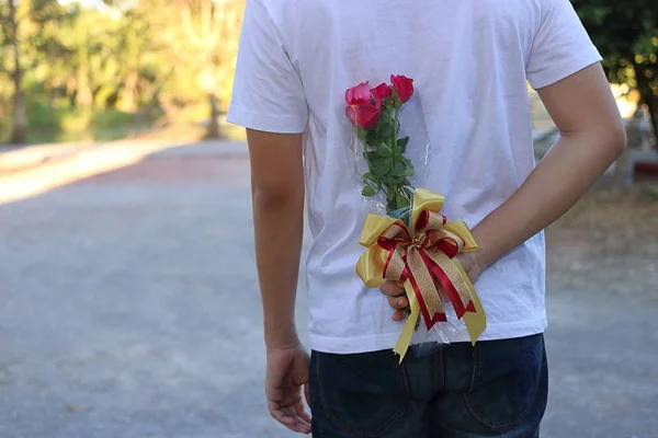 Jovem escondendo rosas vermelhas nas costas para surpreender sua namorada em honra do dia dos namorados — Fotografia de Stock