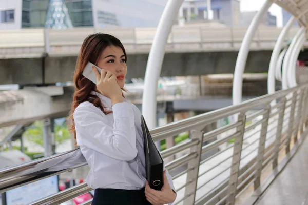 Unga asiatiska affärskvinna prata telefon och hålla dokumentmapp i händerna på urbana staden bakgrund. — Stockfoto