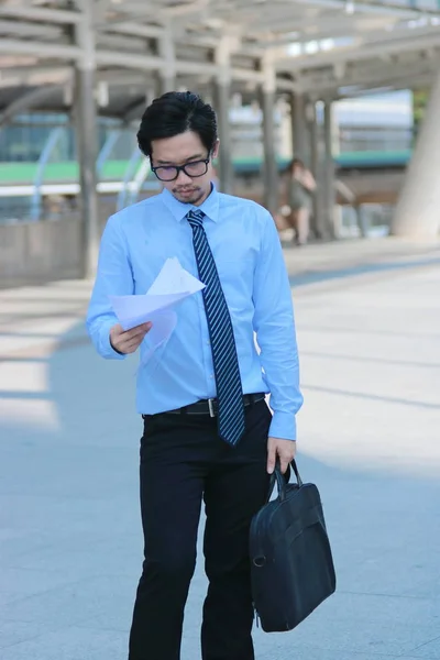 Portrait of young Asian business man standing and looking charts or paperwork at urban building background.