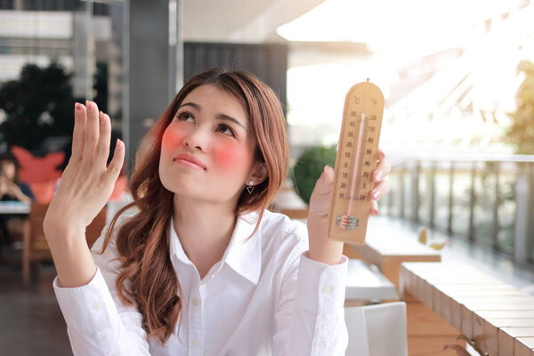 Portrait of young Asian woman holding thermometer and feeling so hot with high temperature on the desk against sunshine effect background.