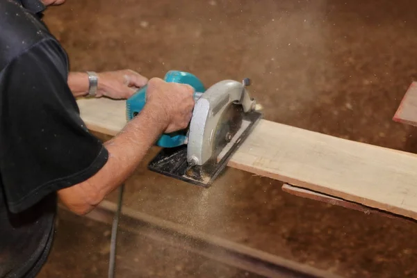 Electric circular saw is being cut a piece of wood against hands of senior carpenter in carpentry workshop. — Stock Photo, Image