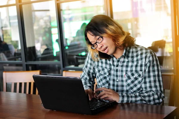 Портрет красивого молодого азиатского сотрудника, разговаривающего по телефону и работающего против своего ноутбука в офисе . — стоковое фото