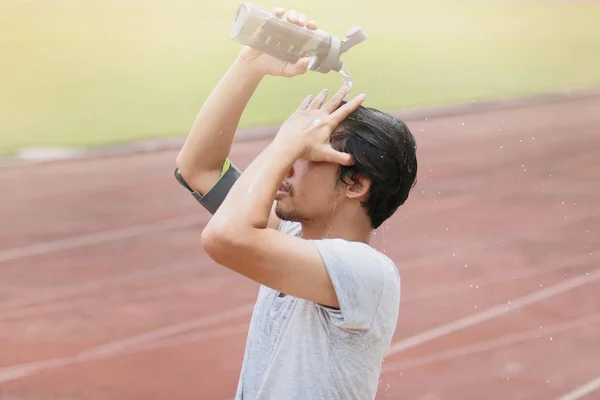 Saludable joven asiático fitness hombre salpicando agua en su cara después de correr en pista de estadio. Efecto sol . — Foto de Stock