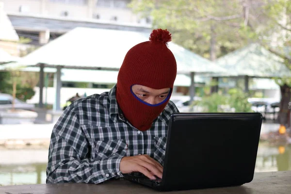 Hacker mascarado em uma balaclava sentado em uma mesa roubando informações com laptop. Conceito de computador criminal . — Fotografia de Stock