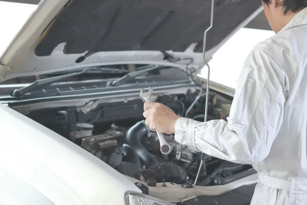Vue latérale du mécanicien automobile en uniforme blanc avec clé diagnostiquant le moteur sous le capot de la voiture au garage de réparation . — Photo
