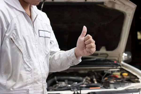 Gros plan mains du jeune mécanicien professionnel montrant pouce vers le haut comme signe de succès avec la voiture dans le capot ouvert à l'arrière-plan du garage. Service de réparation automobile . — Photo