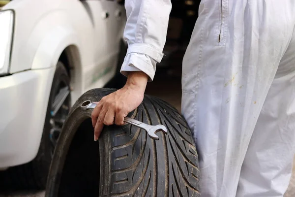 Mãos de mecânico automotivo em uniforme com pneu e chave para a fixação de carro no fundo da garagem de reparação . — Fotografia de Stock