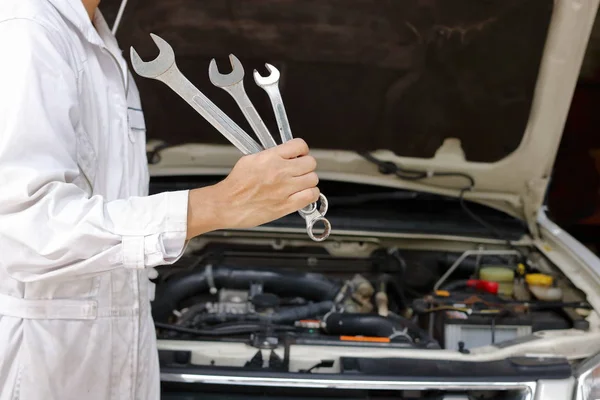 Portrait de jeune mécanicien professionnel en uniforme blanc tenant des clés contre la voiture en capot ouvert au garage de réparation . — Photo