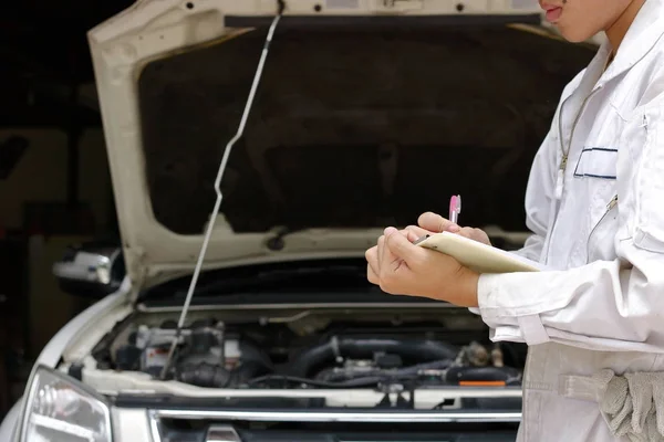 Joven mecánico de automóviles en la escritura uniforme en el portapapeles contra el coche en capucha abierta en el garaje de reparación. Concepto de servicio de mantenimiento . — Foto de Stock