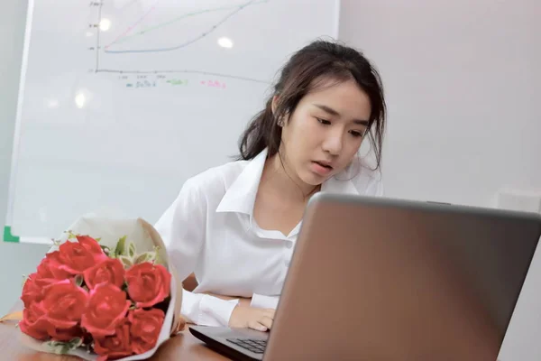 Привлекательная молодая азиатская деловая женщина, работающая с ноутбуком и букет красных роз на столе в офисе . — стоковое фото
