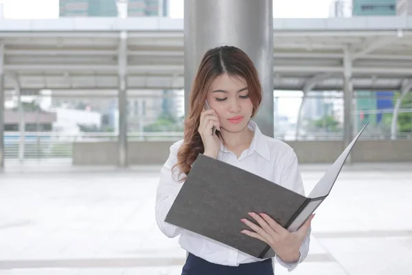 Вид спереди на красивую молодую азиатскую деловую женщину, разговаривающую по телефону и ищущую документы в руках у себя за пределами офиса — стоковое фото