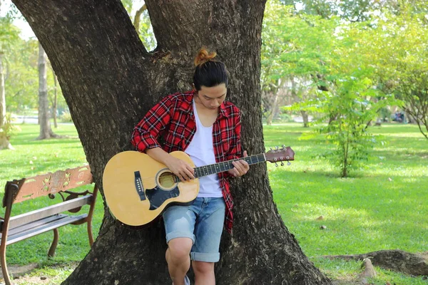 나무 서 있는 아름 다운 자연 배경에서 어쿠스틱 기타를 연주는 젊은 hipster 남자의 초상화. — 스톡 사진