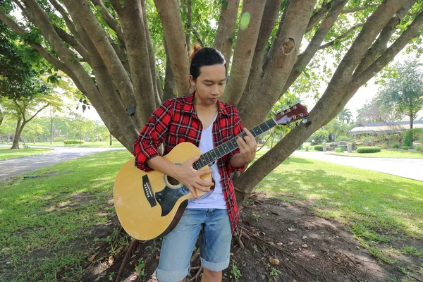 Широкий кутовий знімок молодого хіпстера, який грає музику на акустичній гітарі на красивому фоні природи . — стокове фото
