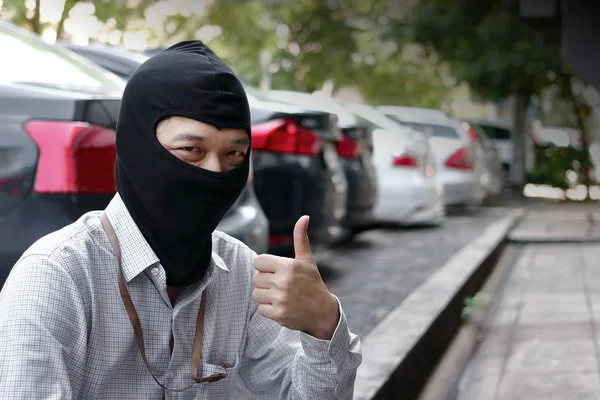 Masked burglar wearing a balaclava showing thumps up  ready to burglary against car background. Insurance crime concept.