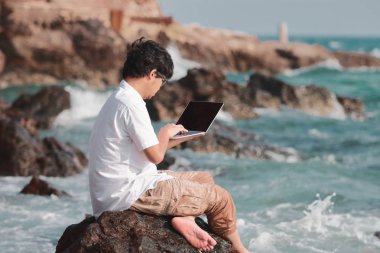 Turkuaz deniz kaya üzerinde oturan dizüstü bilgisayar ile çekici Asyalı gencin arkadan görünüş