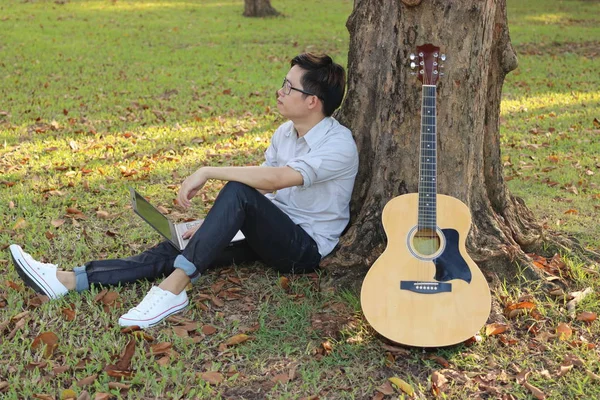 Красивий молодий розслаблений чоловік сидить на зеленій траві і щастя з ноутбуком проти акустичної гітари в парку на відкритому повітрі — стокове фото