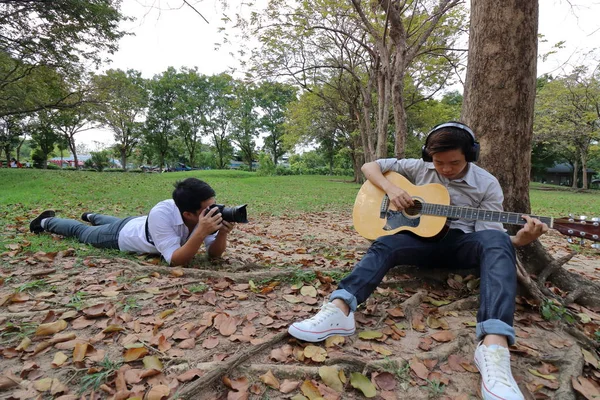 Fotografía de gran angular del fotógrafo tomando una foto de un joven relajado con auriculares tocando la guitarra acústica y sentado debajo de un árbol en el parque al aire libre . — Foto de Stock