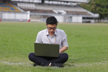 Rahat genç Asyalı iş adamı yeşil çimlerde dizüstü bilgisayarla çalışıyor.