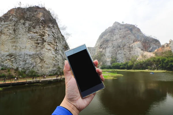 Human hand holding mobile smart phone with scenic view of stone mountain and lake background.