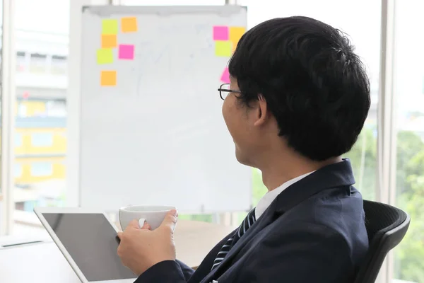 Вид на молодого азиатского бизнесмена, держащего чашку кофе и смотрящего на планшет в руках в офисе . — стоковое фото