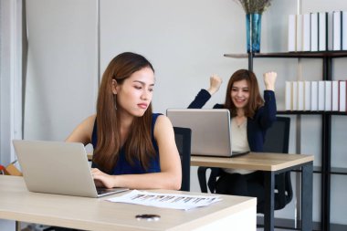 Young angry envious Asian business woman looking successful competitor colleague in office. clipart