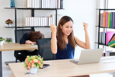 Successful Asian business woman raising hands with competitor colleague bend down head on the desk in office. clipart
