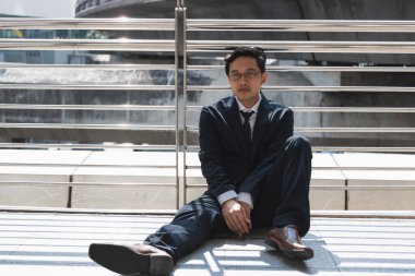 Unemployed stressed young Asian business man sitting on floor outdoors. Failure and layoff concept.
