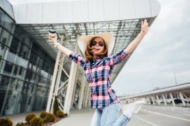 Genç kız öğrenci seyahat tek kullanımlık Kupası holding, atlama ve şapka sallayarak ekose gömlekli. Özgürlük seyahat ve tatil kavramı.