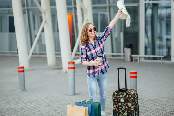 Young hipster woman in sunglasses, plaid shirt with suitcase and disposable cup waving straw for taxi near terminal airport. Freedom Travel and Holiday Concept.