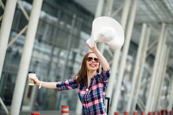 Mujer saltando en gafas de sol con una gran sonrisa en la cara ondeando sombrero en el fondo moderno aeropuerto terminal . — Foto de Stock