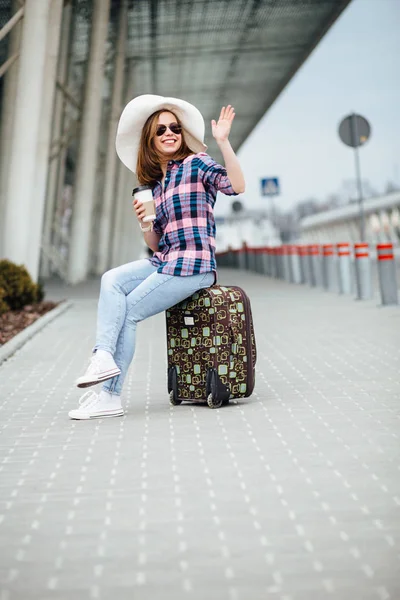 Tourist girl sitting on a suitcase with a disposable cup in her hand. Female traveler dressed in shirt plaid, hat and sunglasses waving hello someonewhile waiting for taxi near aorport terminal. — Stock Photo, Image